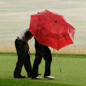 Two golfers holding a red umbrella while rain pours down. 